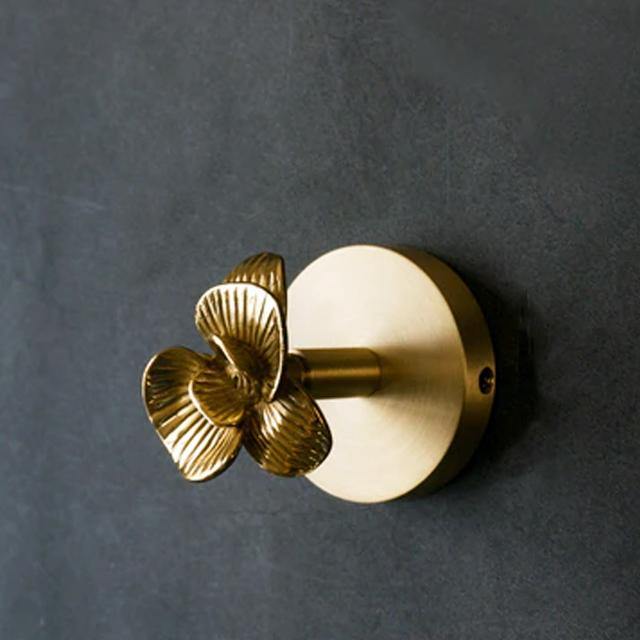 Brass coat hook mounted on a dark grey wall, its round backplate and short peg topped with a flower-shaped casting of ridged petals — the Blume version of the Solid Brass Animal Head Coat Hook.