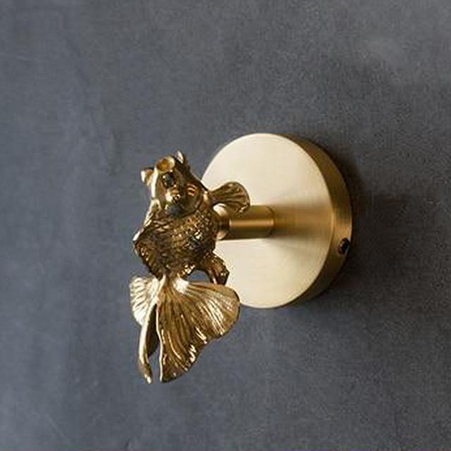 A polished goldfish-shaped solid brass coat hook mounted on a round brass backplate against a dark grey wall, its fanned tail and fins curving downward.