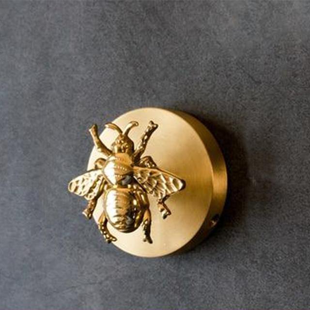 Round polished brass Solid Brass Animal Head Coat Hook with a raised bee design, mounted on a grey textured wall.