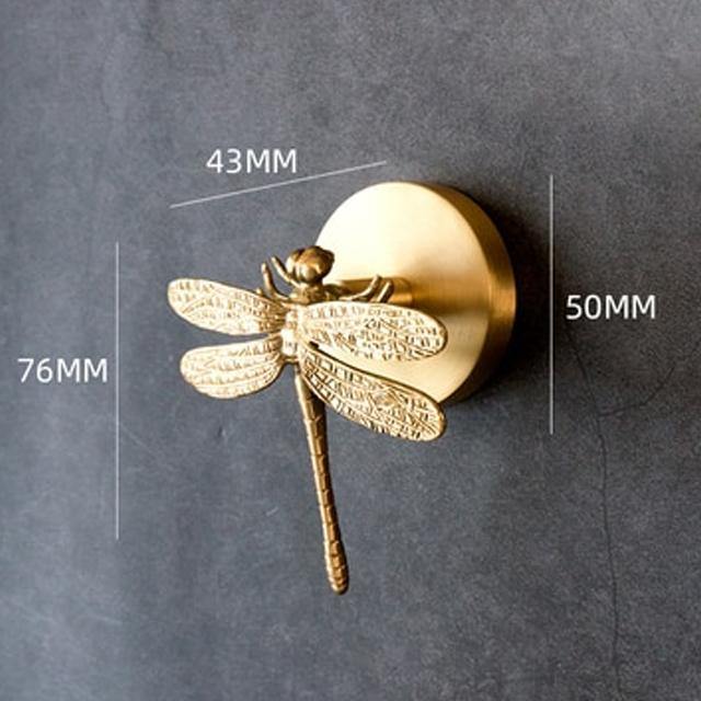 Brass dragonfly coat hook mounted on a dark grey wall, shown with measurement lines marking 43MM across the round wall plate, 50MM for its height and 76MM for the dragonfly's length.