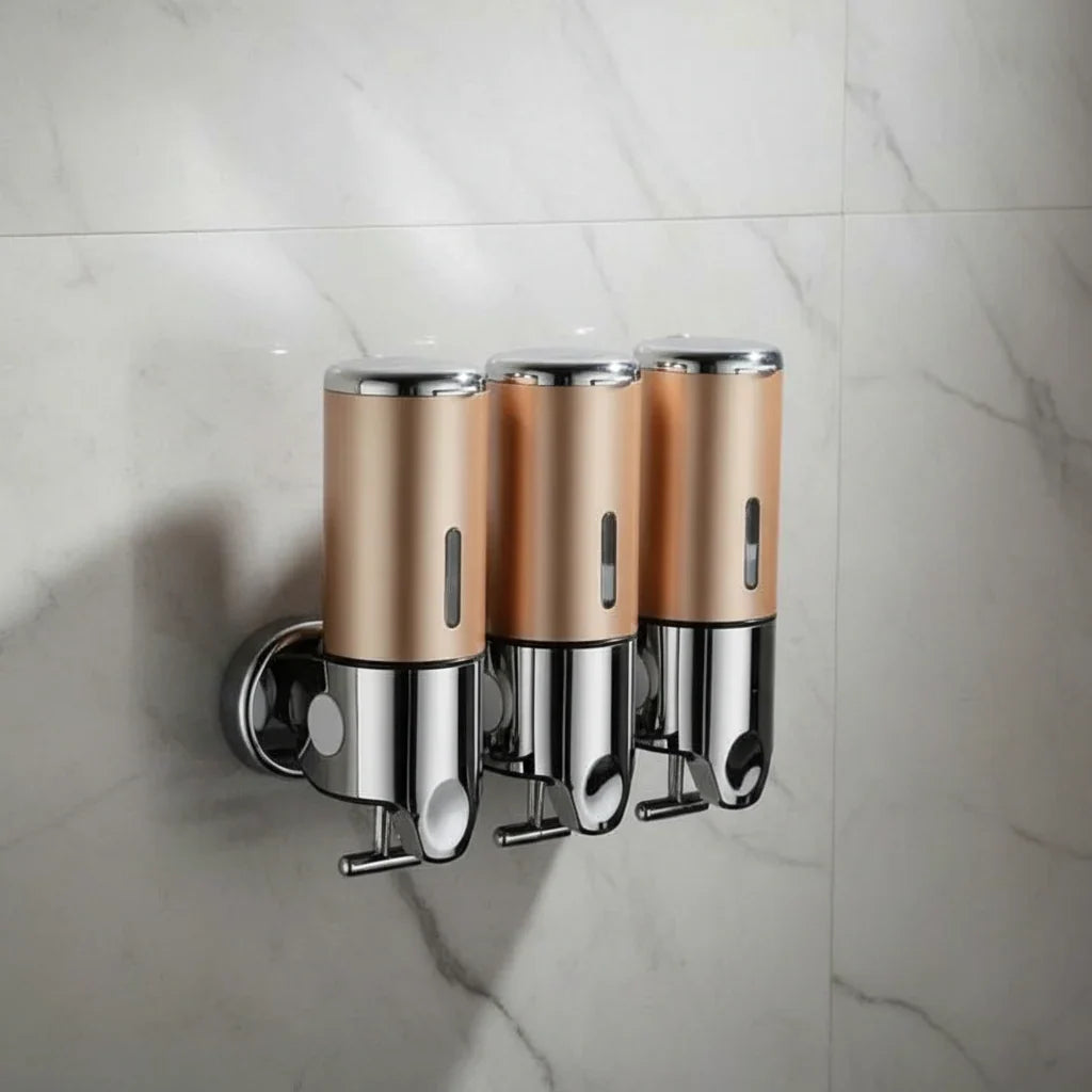 Three gold wall-mounted stainless steel soap dispensers with chrome tops, chrome bases and push levers, fixed side by side on a white marble tile wall.