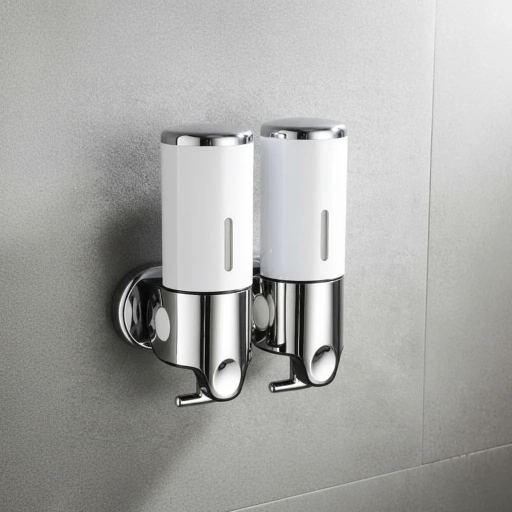 Two white and chrome Wall-Mounted Stainless Steel Soap Dispensers mounted side by side on a grey tiled bathroom wall, each with a push button and a spout underneath.
