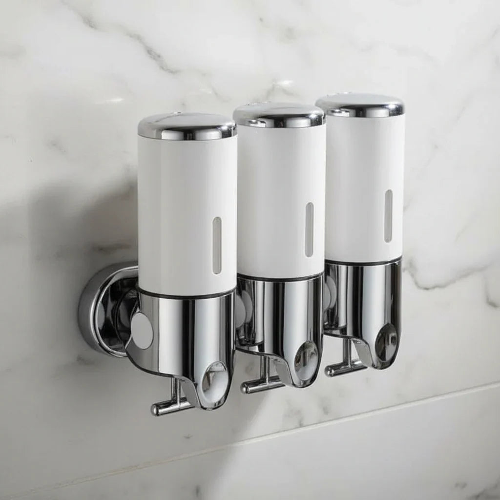Three white and chrome wall-mounted soap dispensers fixed side by side on a white marble bathroom wall, each with a push button on top and a hook beneath the pump spout.