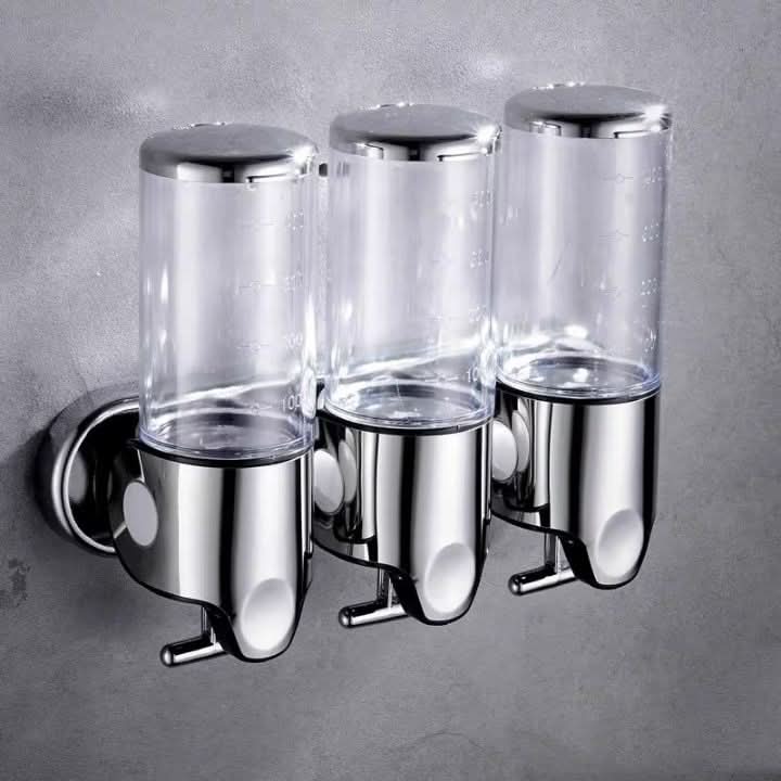 Three wall-mounted stainless steel soap dispensers with clear transparent chambers and chrome pump levers, mounted in a row on a grey concrete wall.