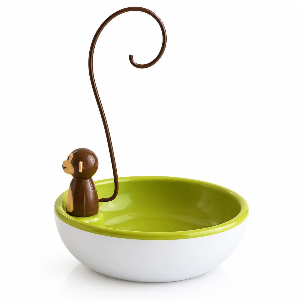 A white ceramic bowl with a bright lime green interior, fitted with a small brown monkey figure on the rim that holds up a tall curved hook arching over the bowl, part of the monkey banana hanger.