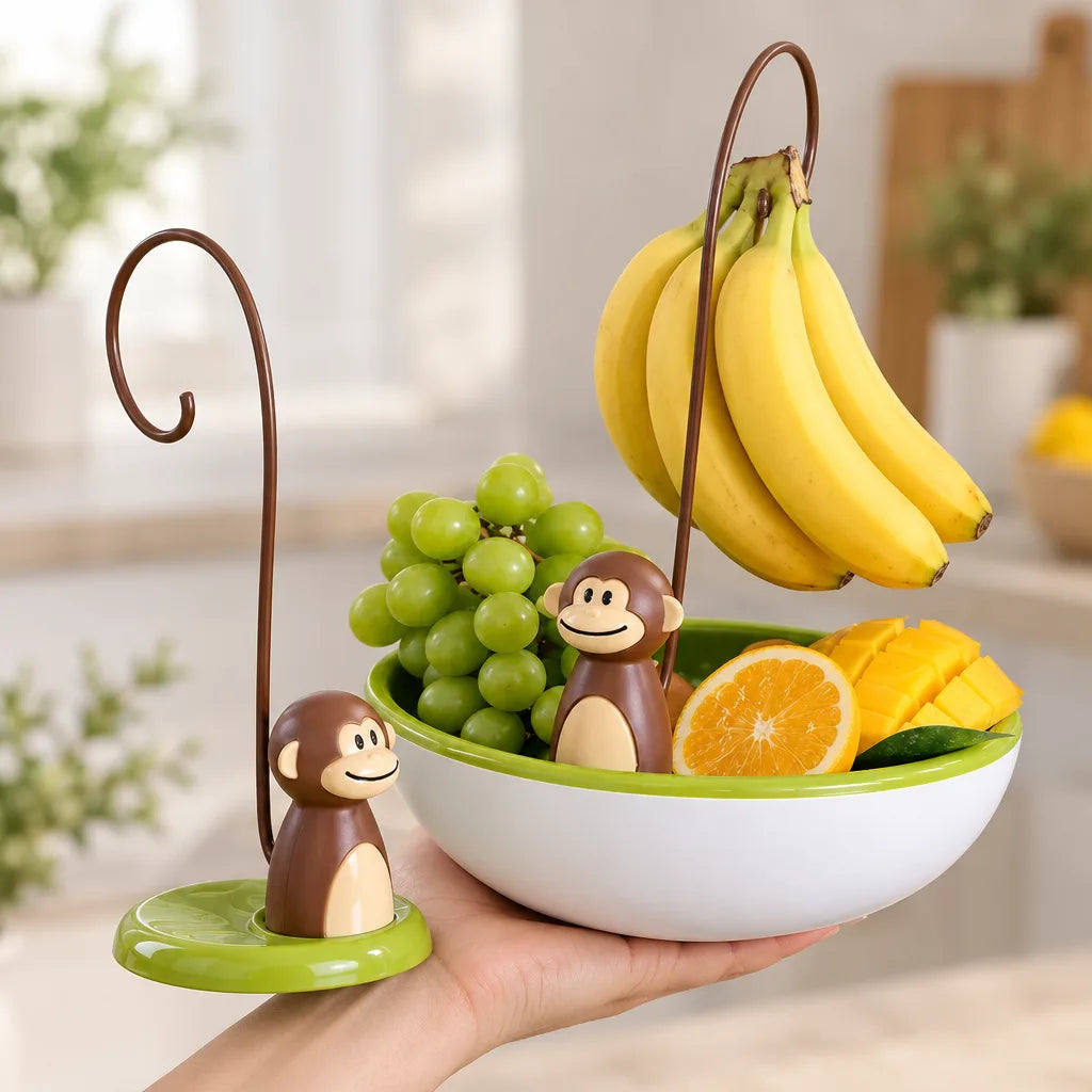 A hand holds a white and green fruit bowl fitted with a monkey banana hanger, its brown curved hook holding a bunch of bananas above green grapes, a halved orange and sliced mango, with a second monkey figure on a matching green base beside it on a bright kitchen counter.