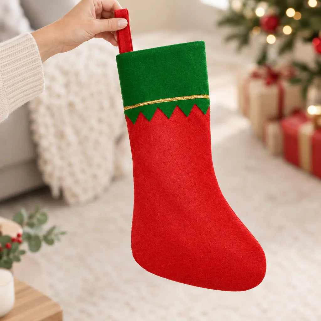 A hand holds up a red felt Christmas Stocking with a green zigzag-edged cuff and gold trim, in a bright living room with a decorated tree and wrapped gifts blurred in the background.