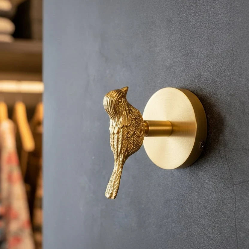Gold bird-shaped Solid Brass Animal Head Coat Hook mounted on a dark grey wall, with a round brass backplate and a clothes rail with hangers blurred in the background.