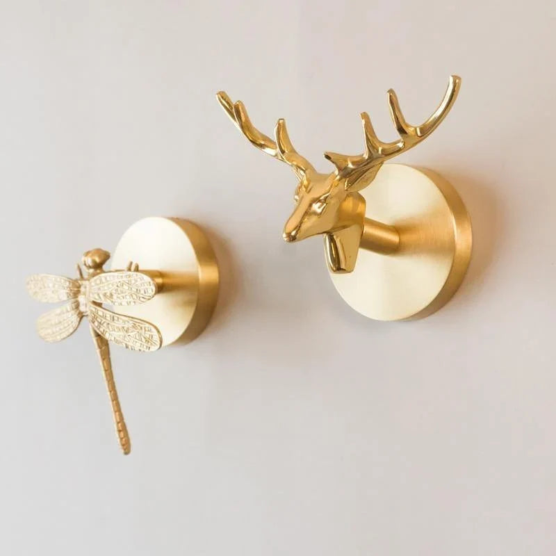 Two gold Solid Brass Animal Head Coat Hooks mounted on a pale wall, one shaped like a dragonfly with detailed wings and one like a stag head with branching antlers, each on a round backplate.