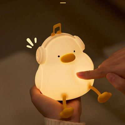 A hand holds the glowing white duck-shaped Squeezable Silicone Night Light while a finger from another hand presses its side, with the duck's orange beak, headphones, feet and a floating music note visible in a dark room.