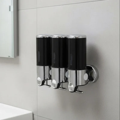 Three black wall-mounted soap dispensers with chrome tops and clear lower chambers, mounted side by side on a light grey tiled bathroom wall beside a mirrored cabinet.