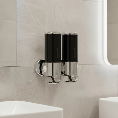 Black double Wall-Mounted Stainless Steel Soap Dispenser with chrome bases and push buttons, mounted on a beige tiled bathroom wall between two white basins.