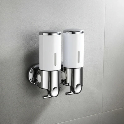 Two white and chrome Wall-Mounted Stainless Steel Soap Dispensers mounted side by side on a grey tiled bathroom wall, each with a push button and a spout underneath.