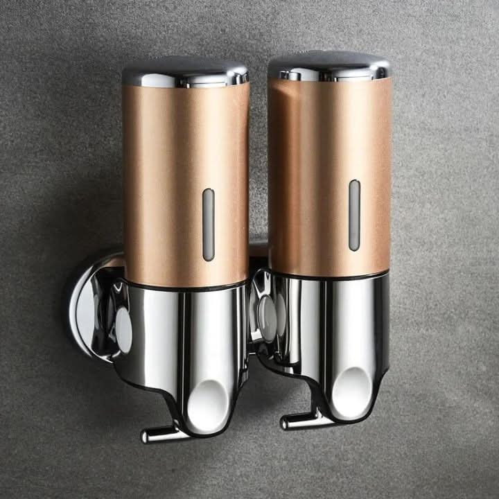 Two gold and chrome Wall-Mounted Stainless Steel Soap Dispenser units mounted side by side on a grey wall, each with a push-down top, a narrow level window and a spout below.