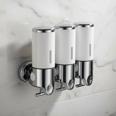 Three white and chrome wall-mounted soap dispensers fixed side by side on a white marble bathroom wall, each with a push button on top and a hook beneath the pump spout.