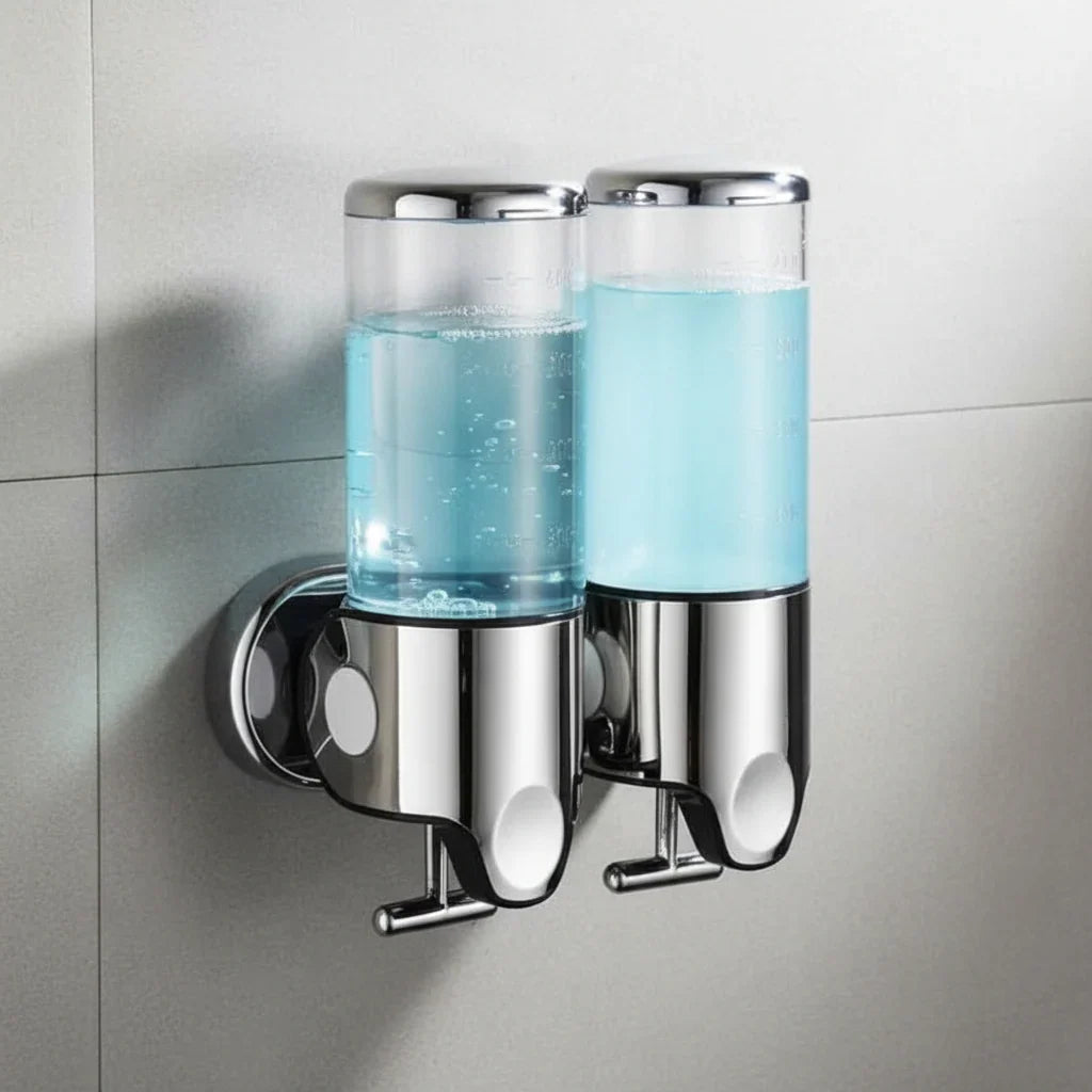 Two transparent Wall-Mounted Stainless Steel Soap Dispenser chambers filled with pale blue liquid, mounted side by side on a grey tiled bathroom wall with chrome caps and push levers.