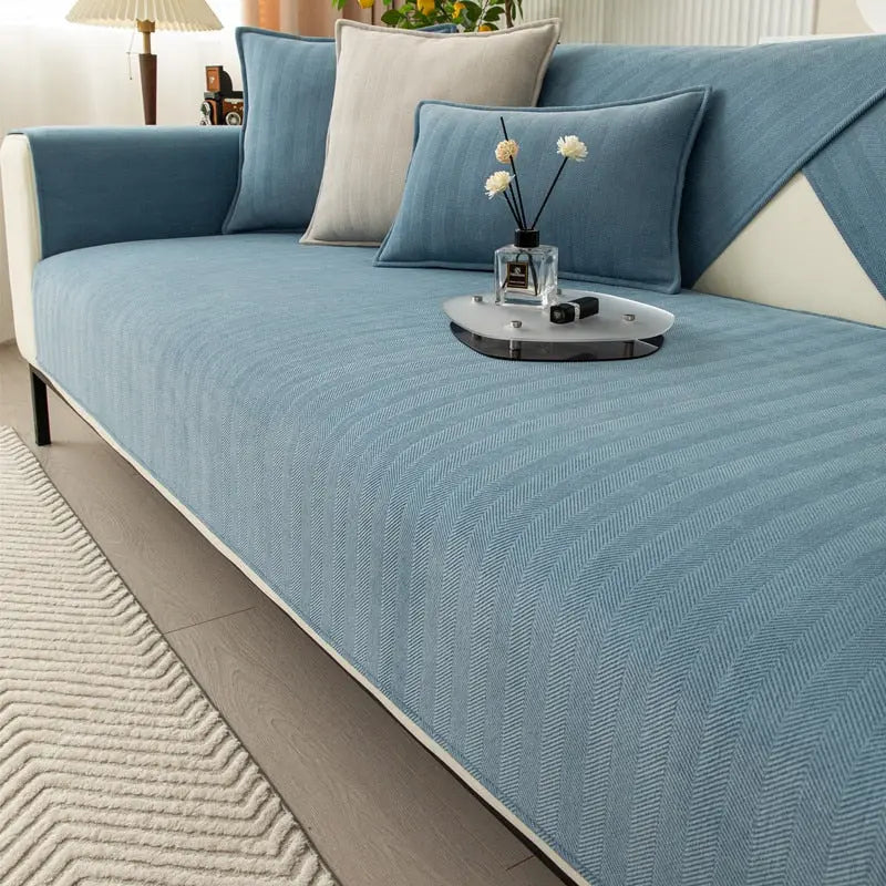 A blue Non-Slip Chenille Sofa Cover with cream trim spread over a sectional sofa, with matching blue cushion covers and a beige pillow against the backrest and a small tray holding a reed diffuser with white flowers on the seat, above a wood floor and a textured cream rug.