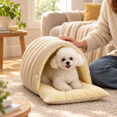 A small white Bichon dog lies smiling inside the beige domed 2-in-1 pet cave bed on a cream rug, while a woman in a knitted sweater kneeling beside it rests her hand on the arched hood above the soft yellow cushion base.