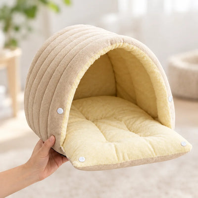 A hand holds up the beige corduroy 2-in-1 cat and dog cave bed, tilted to show its arched dome roof and the soft yellow quilted cushion inside, with white snap buttons along the edges and a bright living room blurred behind.