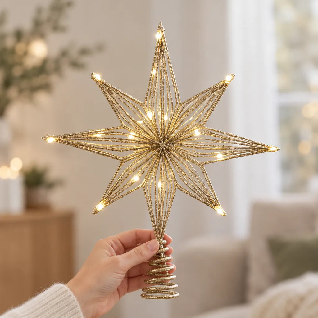 A hand holds up a glittery gold LED star tree topper with eight beaded points lit by small warm white lights, its coiled spring base visible below, in a bright living room with a sofa and window blurred behind.