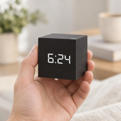 A hand holds a small black cube-shaped digital LED alarm clock showing 6:24 in white digits, with a blurred bedside scene of a potted plant, books and a mug behind it.