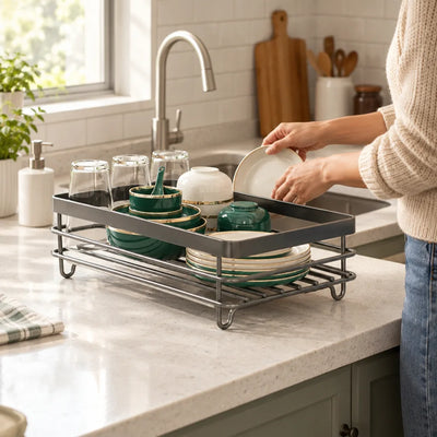 A dish drying rack on a white speckled kitchen counter holds upturned glasses, green and gold bowls and plates, while a woman in a cream sweater places a cream saucer into it beside the sink.