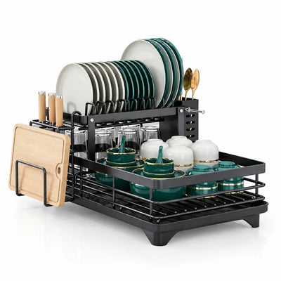 Black two-tier dish drying rack holding white and green plates on the upper shelf, glasses and gold-rimmed green bowls and cups below, with a wooden cutting board and knives in the side holders, gold spoons in the utensil cup, and a drainage tray underneath.