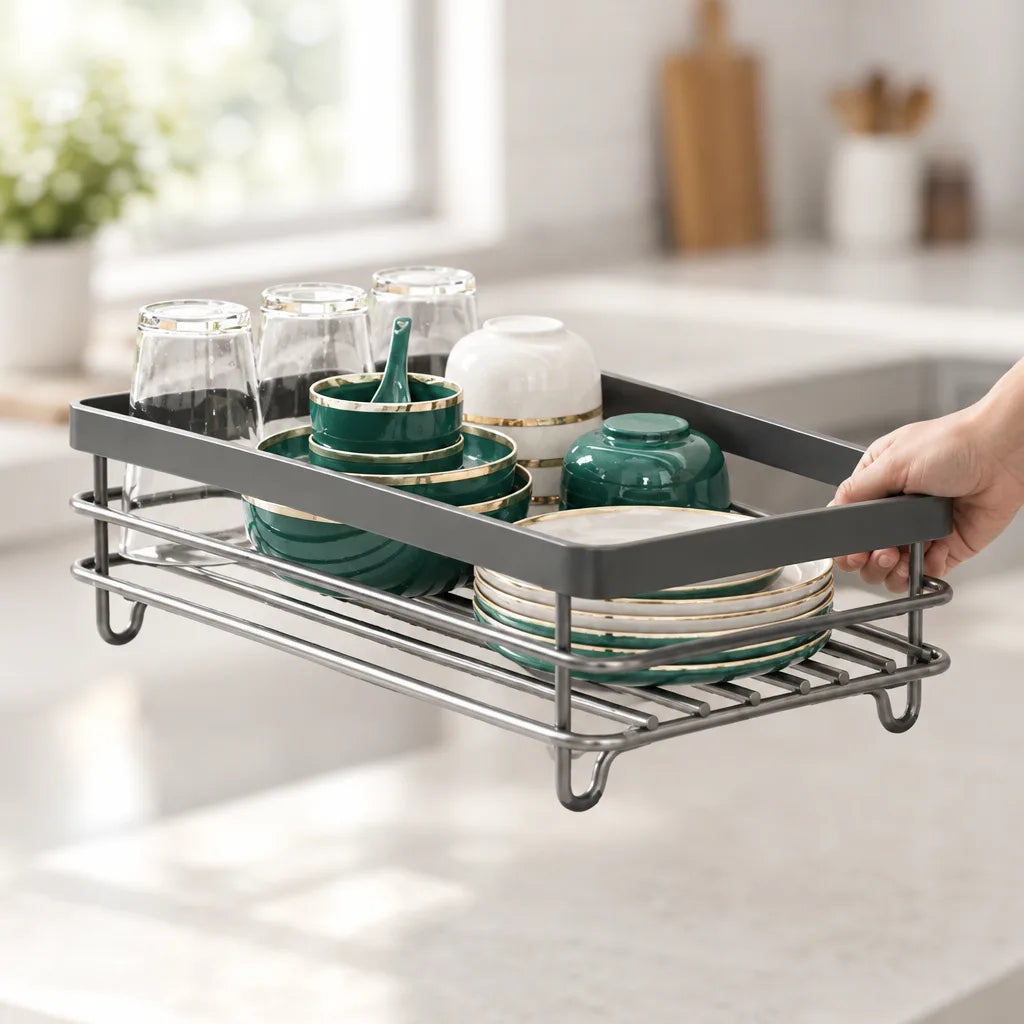 A hand lifts the grey metal dish drying rack off a white kitchen counter, its wire base holding upturned glasses, green and gold-rimmed bowls and a stack of matching plates.