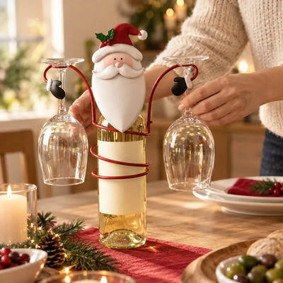 A red metal Santa bottle holder coiled around a bottle of white wine on a festive table, with two wine glasses hanging upside down from its arms as a woman reaches in to lift one, surrounded by pine sprigs, pine cones and candlelight.