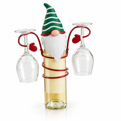 A red metal gnome bottle holder with a striped green hat and white beard sits on a bottle of white wine, its coiled wire arms holding two empty wine glasses upside down on either side against a white background.