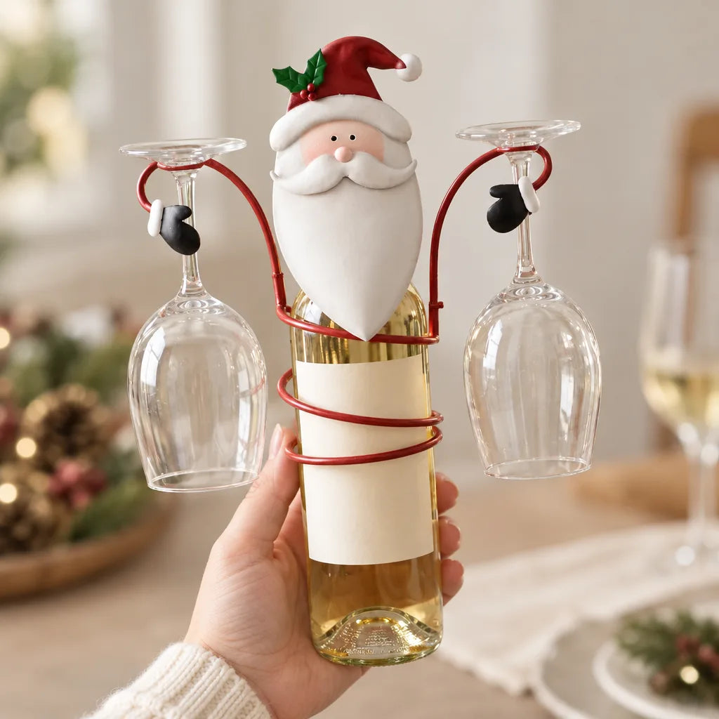 A hand holds a bottle of white wine fitted with the Santa bottle holder, a red metal frame that coils around the bottle, carries a bearded Santa figure at the top and suspends two empty wine glasses upside down by their stems, with a blurred festive table in the background.