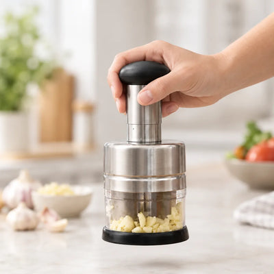 A hand presses down on the black knob of a stainless steel Garlic Chopper on a marble counter, with freshly minced garlic collected in its clear base and whole garlic bulbs nearby.