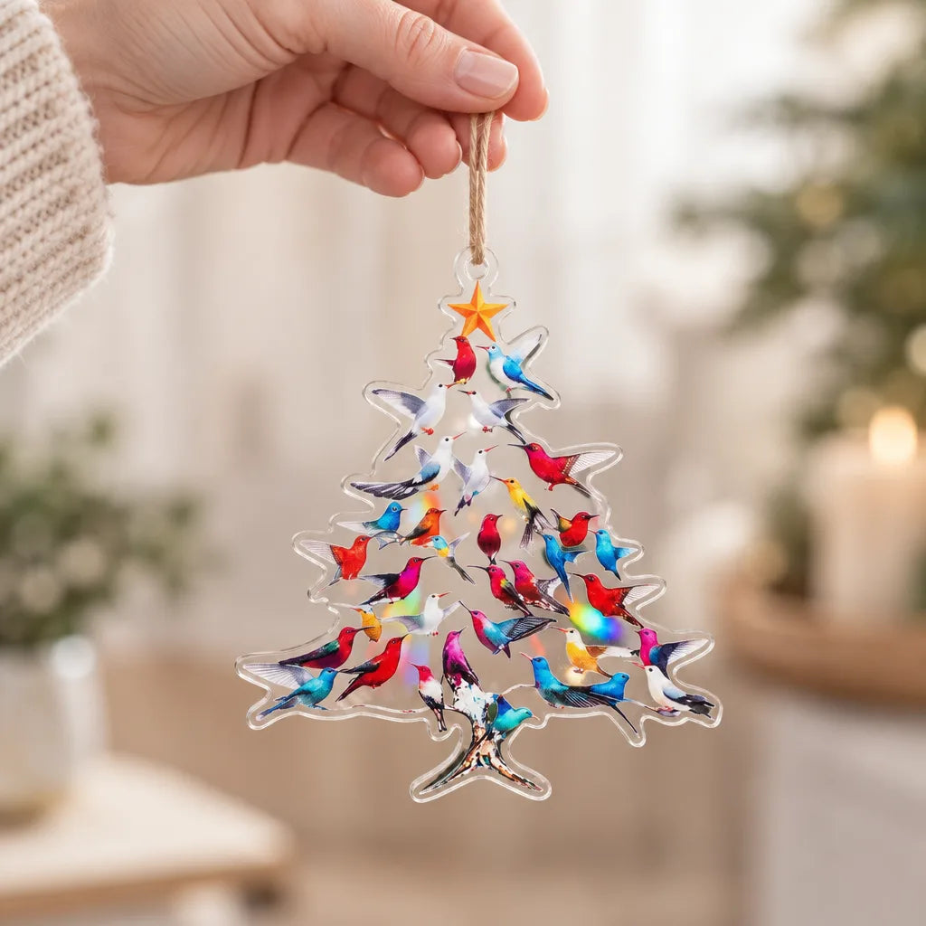 A hand holds a clear acrylic Christmas tree ornament from the Hummingbird Christmas Ornaments set by a twine loop, its tree shape filled with brightly coloured hummingbirds in red, blue, pink and teal beneath a gold star, with a softly blurred living room behind.