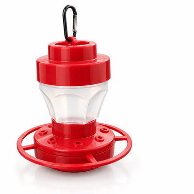 Red hummingbird feeder with a clear tapered reservoir, a wide base with eight flower-shaped feeding ports, a circular perch ring around the rim and a black carabiner clip on top, shown against a plain white background.