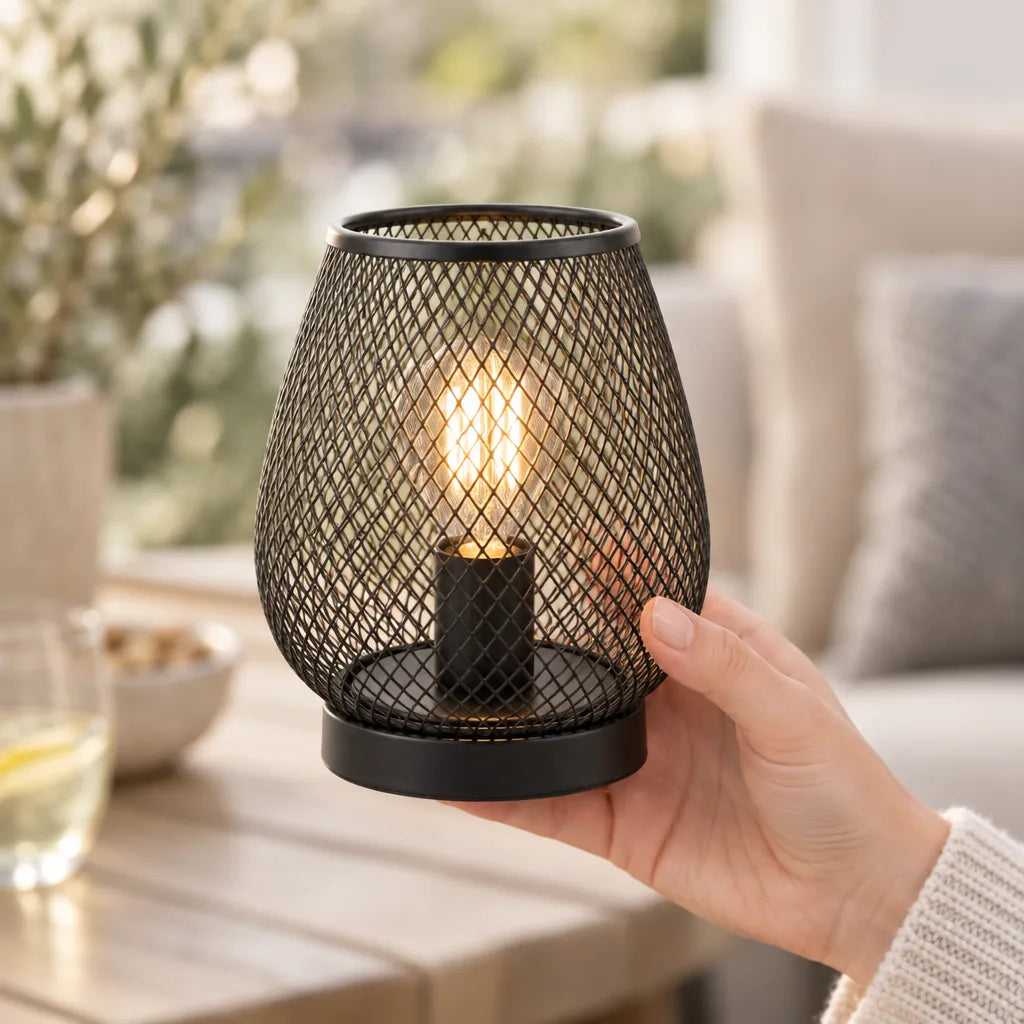 A hand holds the small black mesh LED lantern, its warm filament bulb glowing through the woven metal, above a wooden table set with a glass of white wine and a bowl of nuts beside a cushioned sofa.