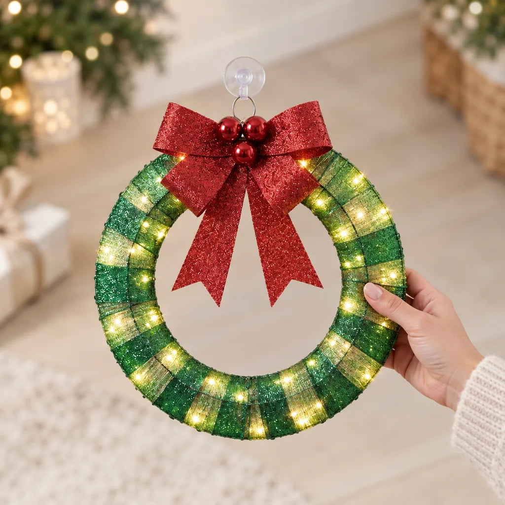 A hand holds up a round LED Christmas wreath wrapped in green and gold glittery ribbon with warm white lights glowing along it, topped by a red glitter bow, three red baubles and a clear suction cup hanger, in front of a softly blurred living room with a lit tree, candles and wrapped gifts.