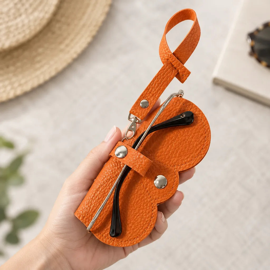 A hand holds an open orange pebbled leather glasses case with a wrist strap and silver snap closures, with a pair of black-armed glasses tucked inside, above a pale woven mat with a straw hat and notebook blurred in the background.