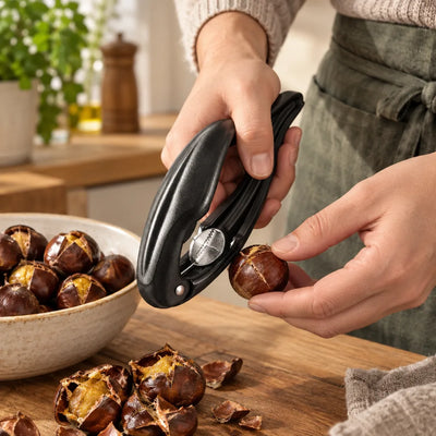 Hands in a grey apron using a black nutcracker for chestnuts to score a chestnut shell at a wooden kitchen counter, with a bowl of split chestnuts and loose shells nearby.