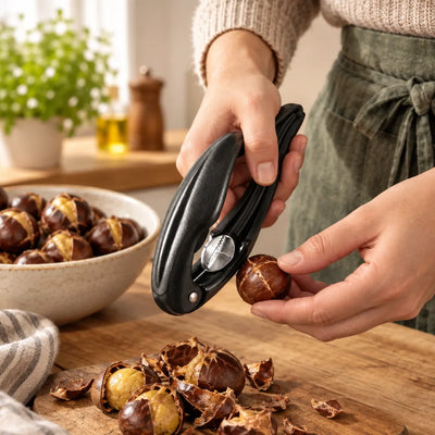 Close-up of a person in a green apron holding the black Nutcracker for Chestnuts in one hand and a scored chestnut in the other, over a wooden board scattered with cracked shells and peeled chestnuts, with a bowl of scored chestnuts nearby.