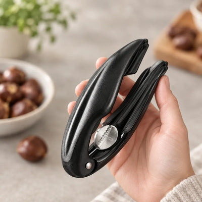 A hand holds the black Nutcracker for Chestnuts, its hinged handles open to show the serrated stainless steel cracking jaw, with a bowl of chestnuts blurred in the background.