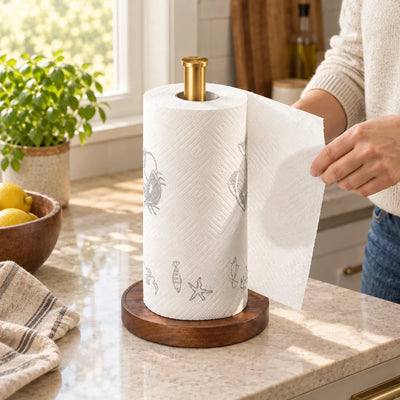 A walnut paper towel holder with a brushed gold post stands on a speckled kitchen counter, holding a roll printed with faint crab, fish and starfish drawings, as a woman in a cream sweater tears off a sheet; a basil plant, a wooden bowl of lemons and a striped tea towel sit nearby in the sunlit kitchen.