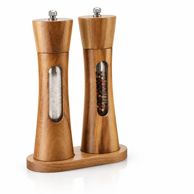 Two tapered acacia wood salt and pepper grinders with stainless steel knobs standing in a matching oval wooden base, each with a narrow window showing coarse white salt in one and mixed peppercorns in the other.