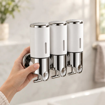 A hand holds a triple white and chrome Wall-Mounted Soap Dispenser against a marble bathroom wall, with each of the three chambers showing a narrow level window and a pump lever below.