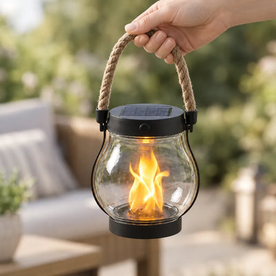 A hand holds a solar flame lantern by its rope handle on a sunny patio, the clear glass jar glowing with an orange flickering flame effect beneath a black lid with a small solar panel, with blurred garden foliage and outdoor seating behind it.