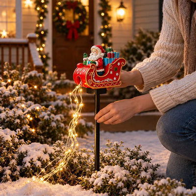 A woman in a cream knit sweater and jeans kneels in a snowy front garden to push the stake of a Santa in sleigh solar garden stake into the ground, its red sleigh loaded with gifts and a trailing tail of warm white fairy lights glowing against the snow-dusted shrubs, with a wreath-hung porch lit behind her.