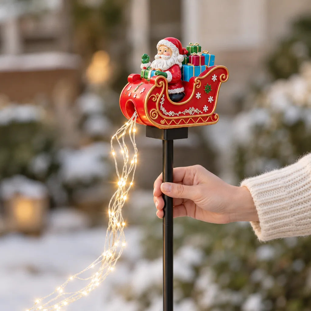 A hand in a cream knit sweater holds the black stake of a Santa in sleigh solar garden stake upright in a snowy yard, its red sleigh loaded with colorful gifts and a trailing strand of warm white fairy lights glowing below.