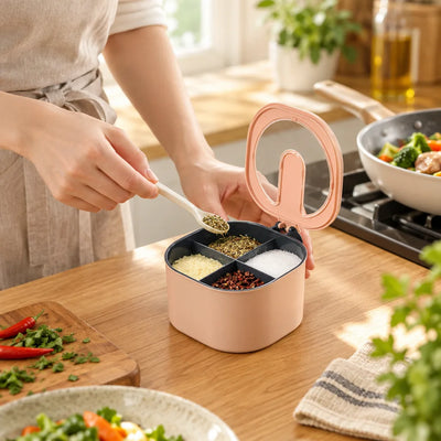 A person in a linen apron lifts a spoonful of dried herbs from a pink four-compartment spice storage jar on a wooden kitchen counter, holding its clear-topped lid in the other hand while the sections below hold herbs, salt, chili flakes and powder, with a pan of stir-fried vegetables on the stove behind.