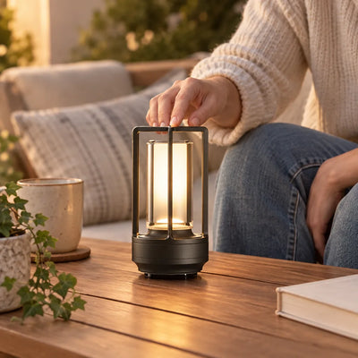 A person in a cream knit sweater and jeans reaches down to touch the top of a small black cordless table lamp glowing warm on a wooden outdoor table, with a ceramic mug, a trailing ivy plant and a closed book nearby.
