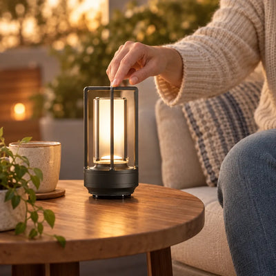 A hand reaches down to touch the top of a small black cordless table lamp glowing warm amber on a round wooden side table, beside a speckled ceramic pot of greenery, with a person in a cream knit sweater seated on a cushioned sofa in the softly lit background.