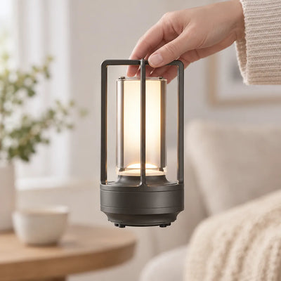 A hand lifts a small dark metal cordless table lamp by its square carry handle, its frosted cylinder glowing warm amber, in a bright living room with a sofa, knit throw, side table and plant blurred behind.